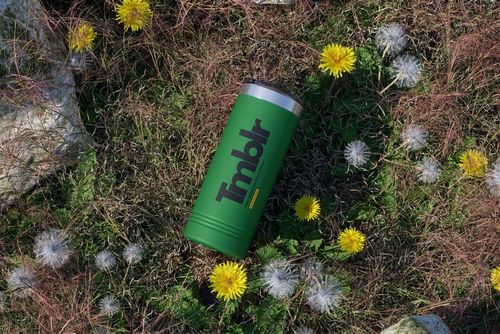 Powder coated skinny tumbler mockup in green placed outdoors on grassy ground with dandelions and stones, top view.