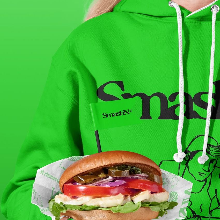 Packaging mockup showing a person in a bright green hoodie holding a vegan burger with branded paper and a small green flag.