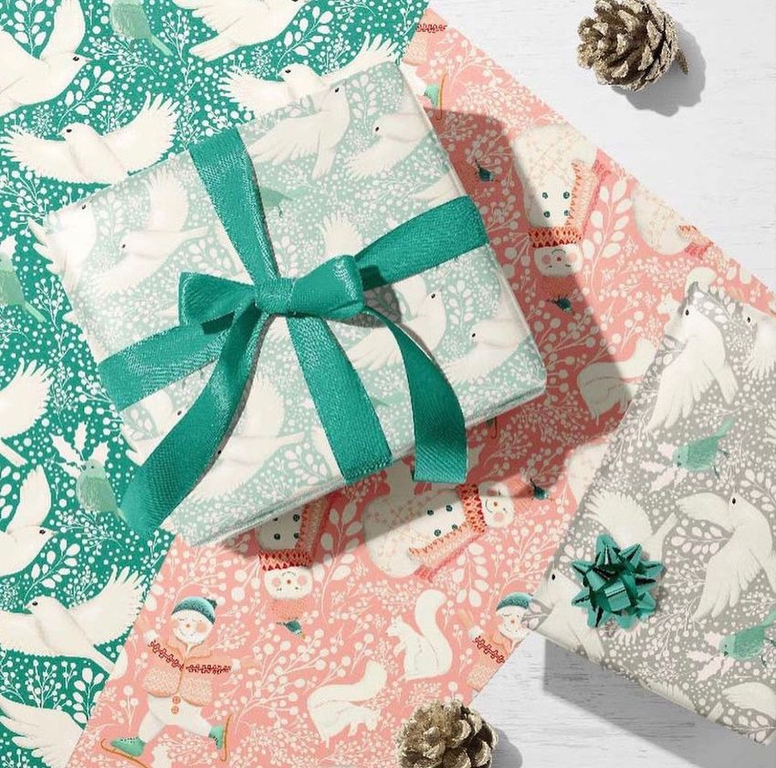 Wrapping paper mockup featuring two wrapped gift boxes with teal bows on patterned paper sheets with birds and snow people designs.
