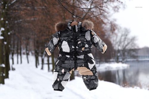 Baby snow suit mockup with fur trim, displayed on a hanger among snowy trees and a frozen lake in the background.
