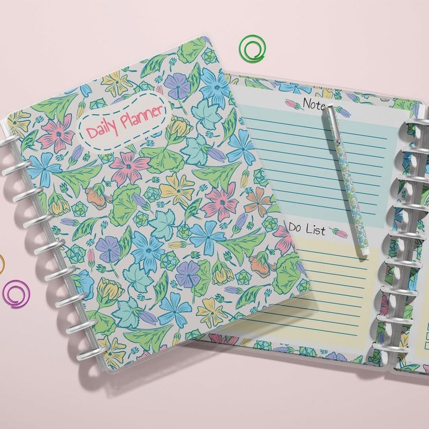 Top view of two spiral planners with colorful tropical floral covers, one closed and one open showing lined pages and a pen.