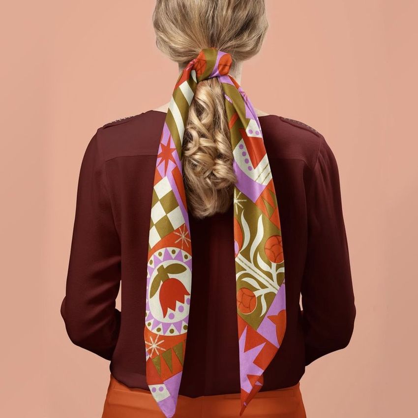 Silk scarf mockup featuring a woman with blonde hair tied in a ponytail, showcasing a colorful patterned scarf against a peach background.