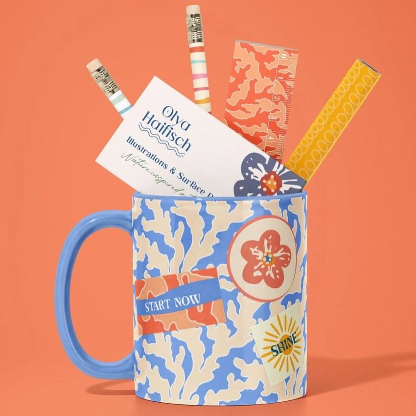 Mug mockup with a blue handle and abstract floral pattern, filled with pencils, a ruler, and business cards against an orange background.