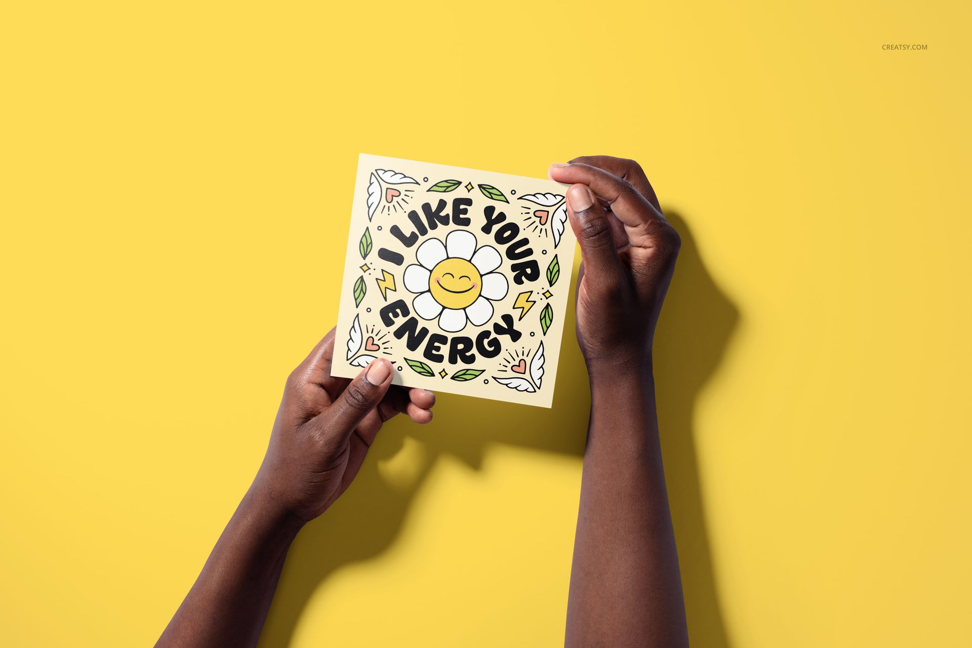 Person holding a square greeting card mockup featuring a smiling flower and colorful doodles, set against a bright yellow surface.