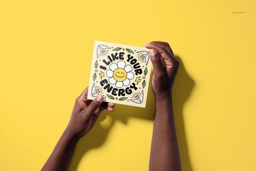 Person holding a square greeting card mockup featuring a smiling flower and colorful doodles, set against a bright yellow surface.