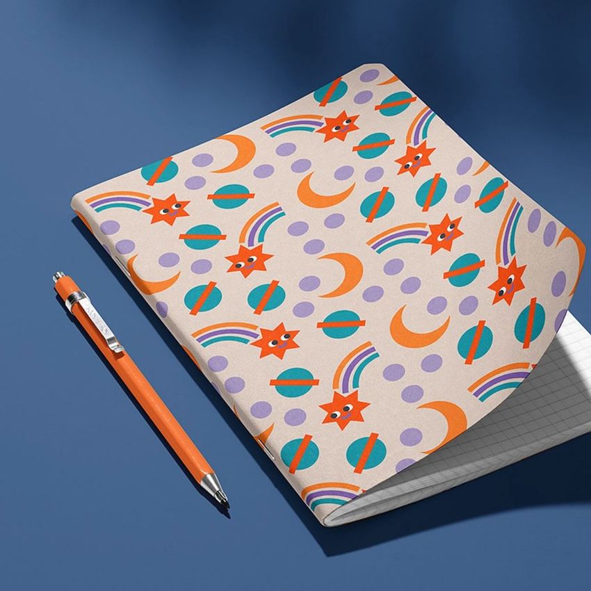 Notebook mockup with a raised corner showing a colorful cover featuring orange stars, moons, rainbows, and circles on a beige background.