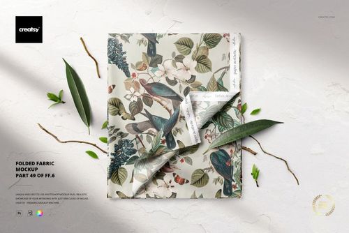 PSD mockup of folded fabric featuring a colorful floral and bird design, shown from a top-down perspective with leaf props.