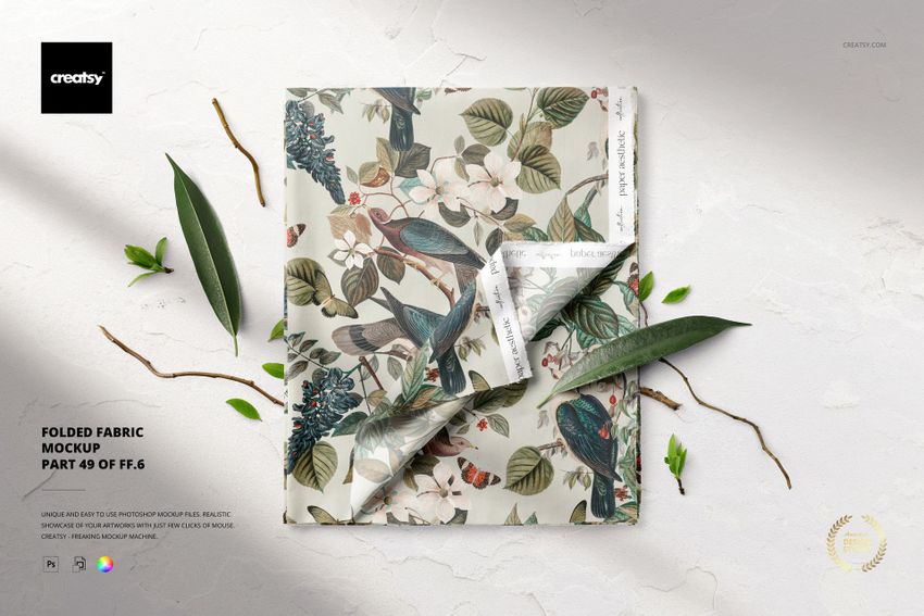 PSD mockup of folded fabric featuring a colorful floral and bird design, shown from a top-down perspective with leaf props.