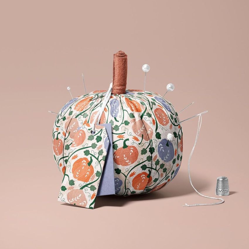 Fabric pumpkin mockup with a colorful pumpkin pattern, used as a pincushion with sewing pins and a thimble on a beige background.