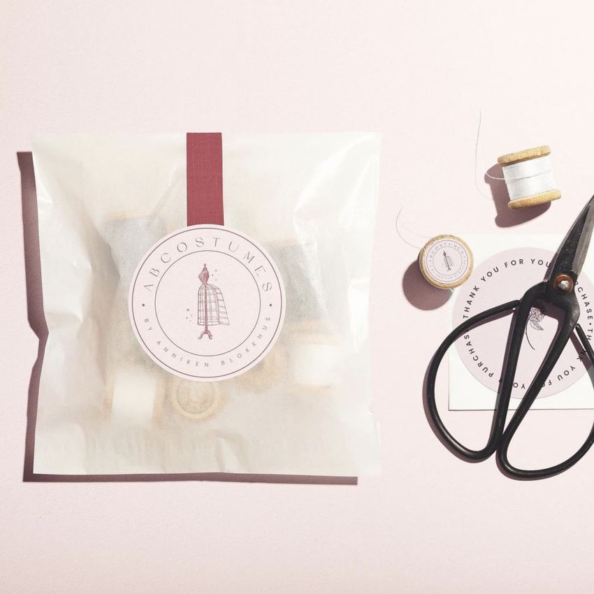 Sticker mockup showing a translucent packaging bag sealed with a round label, surrounded by scissors, thread spools, and a sticker sheet on a pale pink surface.