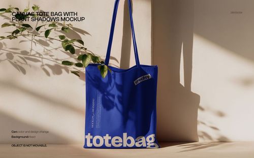 Blue canvas tote bag mockup hanging against a light wall with plant shadows cast across the surface.