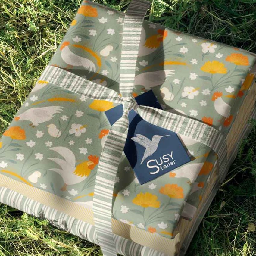 Spring fabric stack mockup showing folded fabrics with chicken and floral patterns, tied with a striped ribbon on grass.
