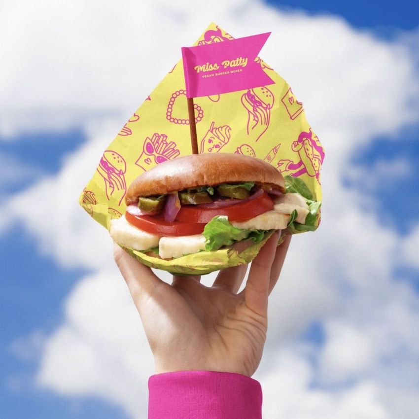 Vegan burger bar packaging mockup featuring a colorful burger and patterned wrap, held up in front of a cloudy sky.