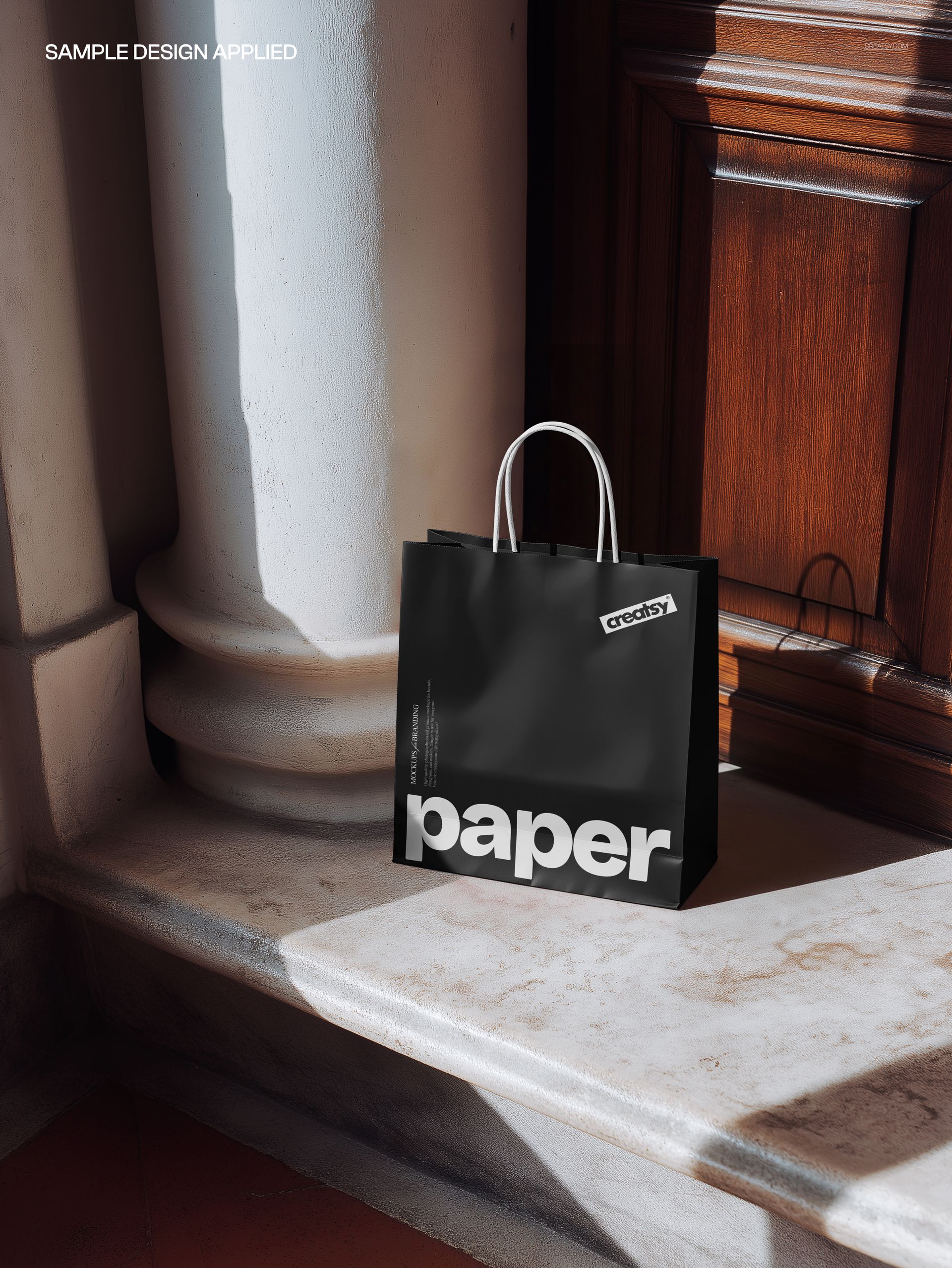 Black paper shopping bag with white handles on marble stairs, sunlight casting shadows, front view mockup.