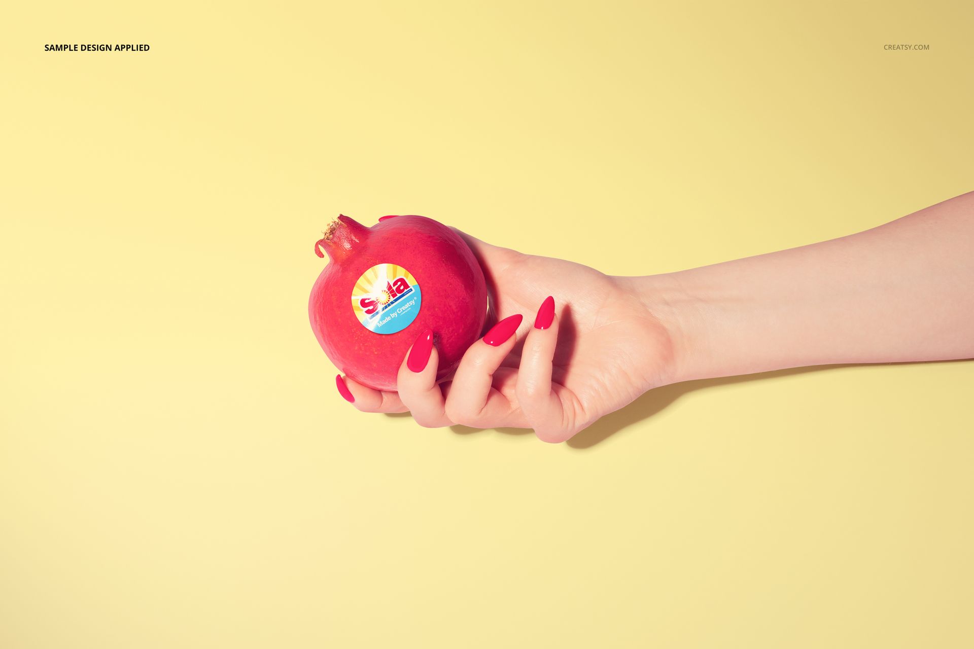 Round fruit sticker mockup displayed on a red pomegranate, held by a hand, with a soft yellow background.