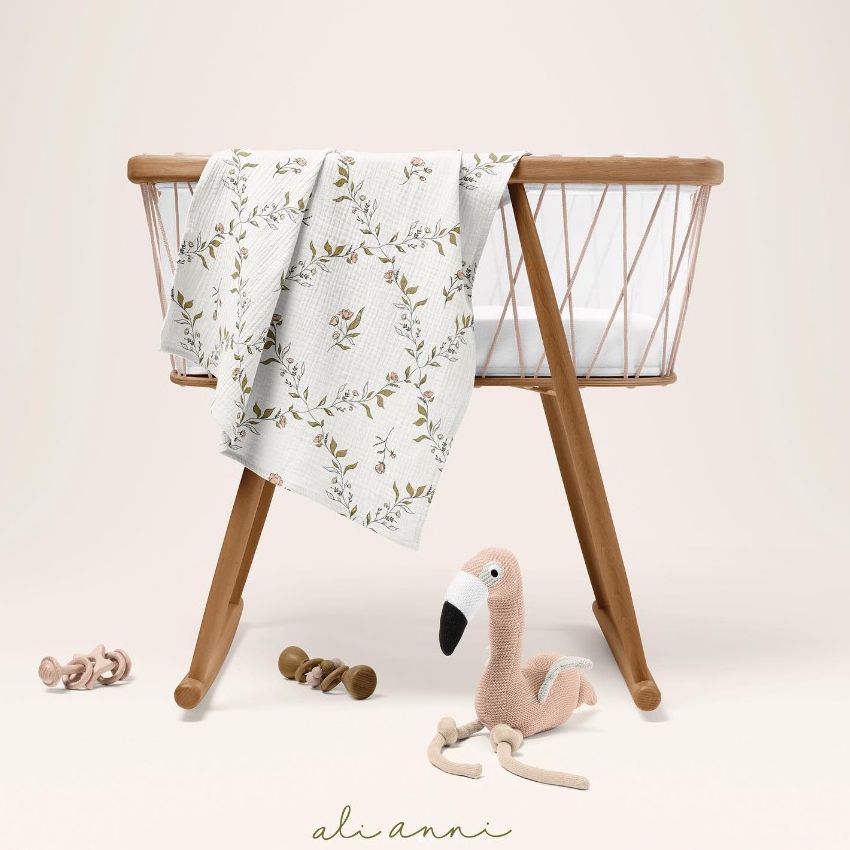 Crinkle muslin swaddle blanket mockup draped over a wooden baby bassinet, with neutral toys and a plush flamingo on the floor.