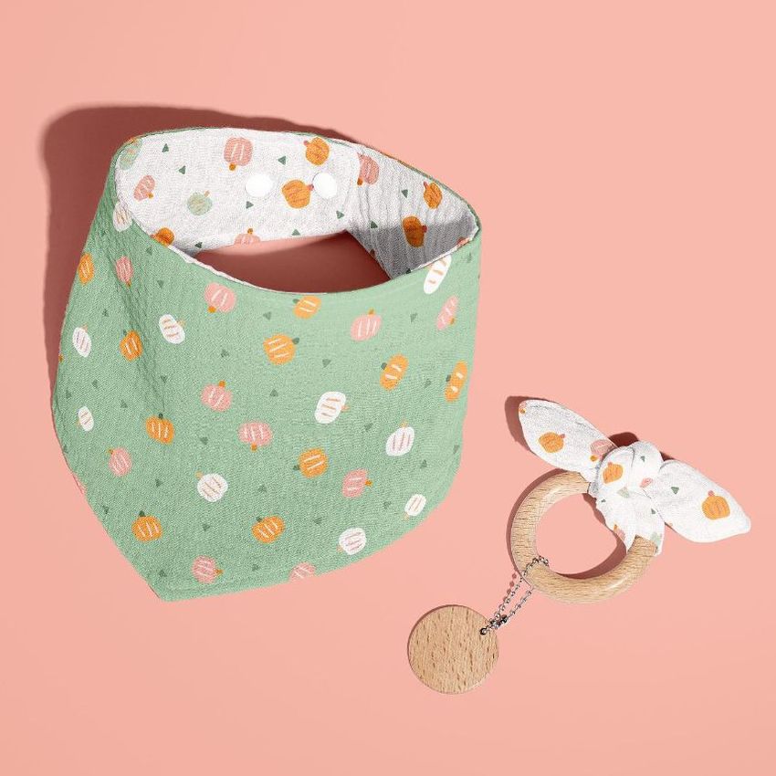 Baby muslin bandana mockup with a green fabric featuring a pumpkin pattern, shown next to a wooden teething ring on a pink background.
