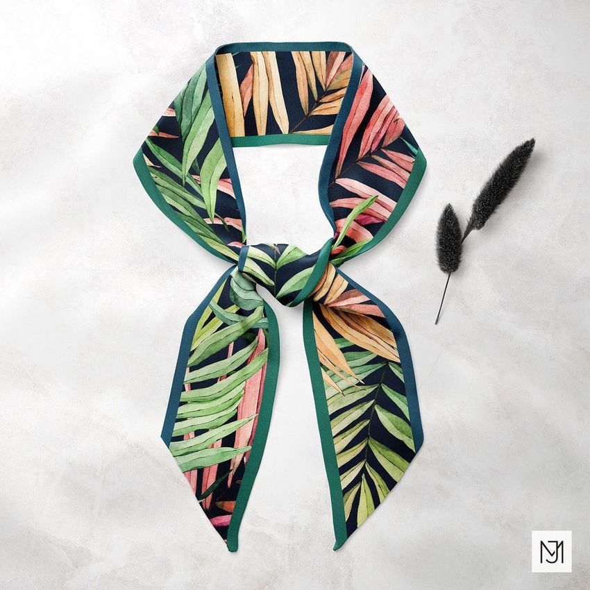 Top view of a tied twilly scarf mockup featuring a tropical leaf pattern in green, orange, and yellow on a light surface.