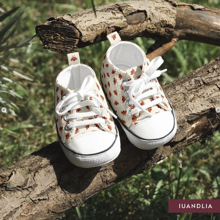 Baby sneakers mockup featuring white shoes with a ladybug pattern, displayed outdoors on a tree branch with grass in the background.