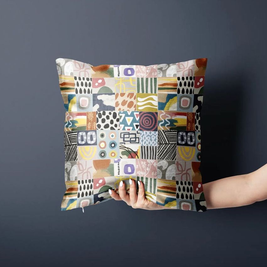 Polyester cushion cover mockup featuring a hand holding a square pillow with a colorful abstract patchwork pattern against a dark wall.