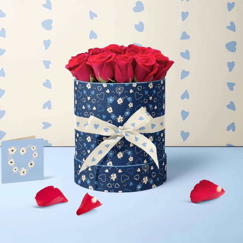 Floral box mockup featuring a round navy blue box with white and yellow flower patterns, filled with red roses and tied with a heart-patterned ribbon, set against a light background with blue hearts.