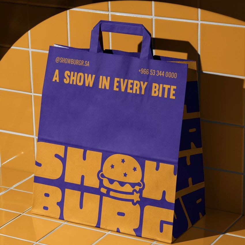 Fast food takeaway paper bag mockup featuring bold purple and orange branding with burger icon and text, set on tiled surface.