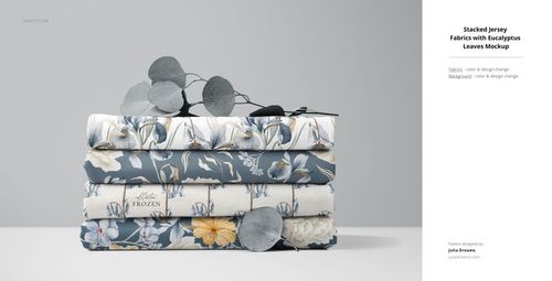 PSD mockup showing stacked jersey textiles featuring floral motifs, displayed with silver eucalyptus leaves on a gray surface.