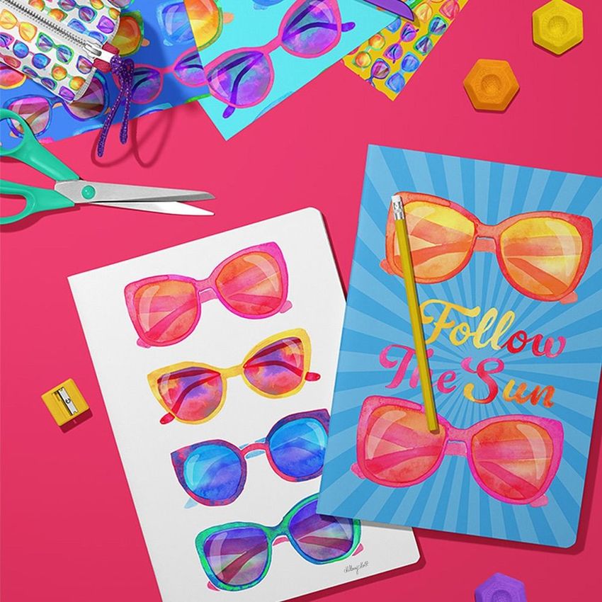 Stationery mockup scene with two A4 notebooks featuring colorful sunglasses designs on a pink surface, surrounded by art supplies.