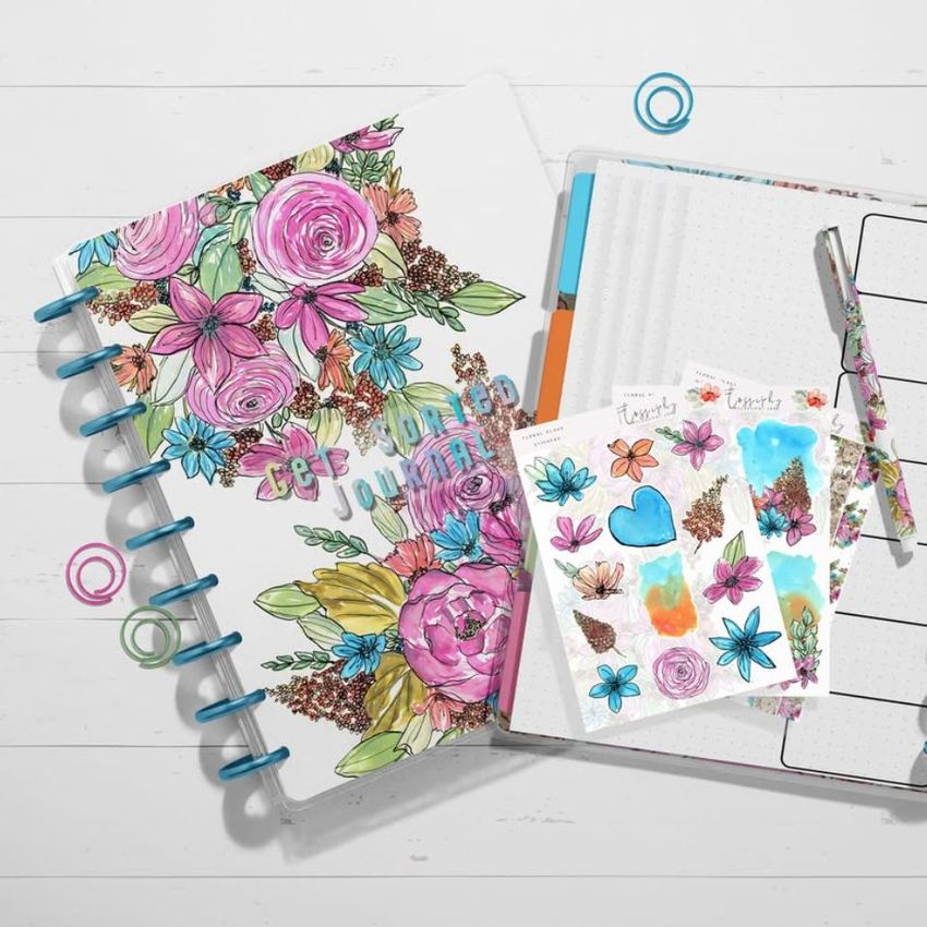 Top view of two spiral-bound planner mockups on a white wooden surface, one with colorful floral cover art and the other open to a dotted page with stickers and a pen.
