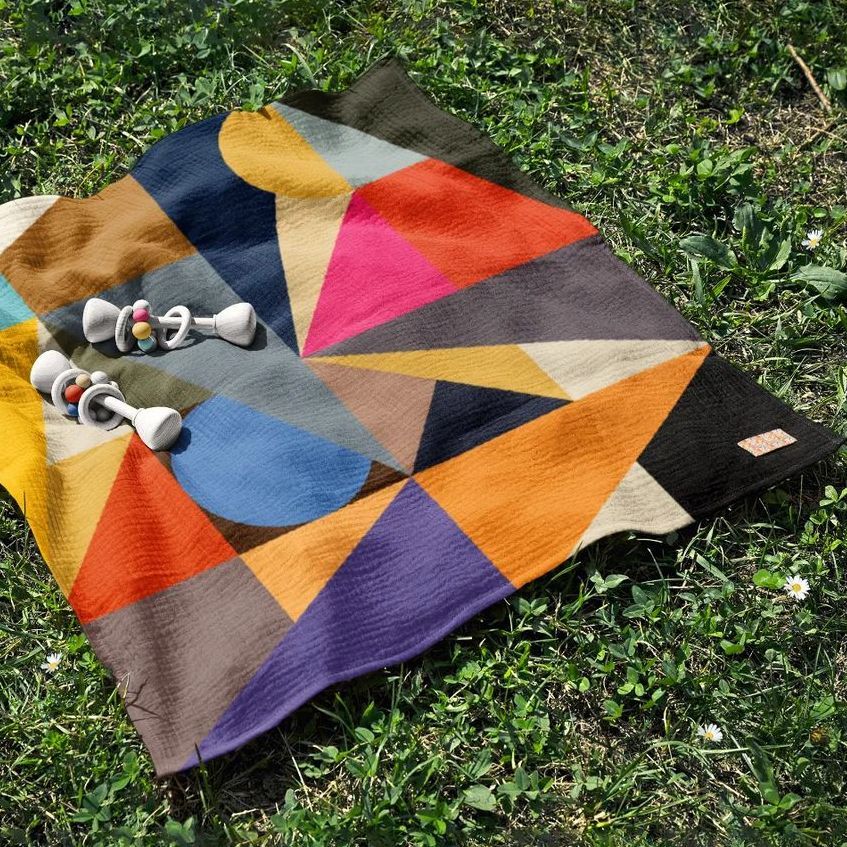Crinkle cotton muslin swaddle blanket mockup with bold geometric shapes in various colors, placed on green grass outdoors.