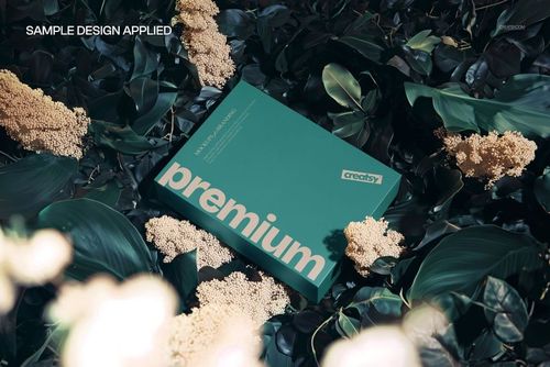 Top view of a green packaging box mockup surrounded by dense botanical greenery and cream-colored blossoms.