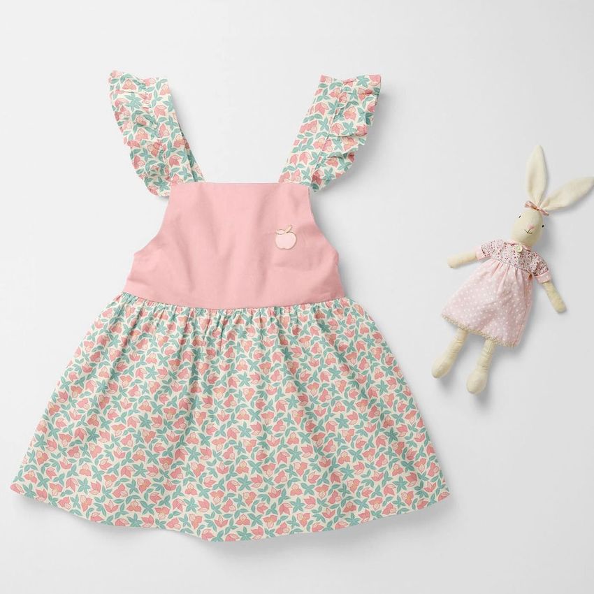 Baby dress mockup featuring a sleeveless dress with pink bodice and floral patterned skirt, displayed on a light surface next to a fabric bunny doll.