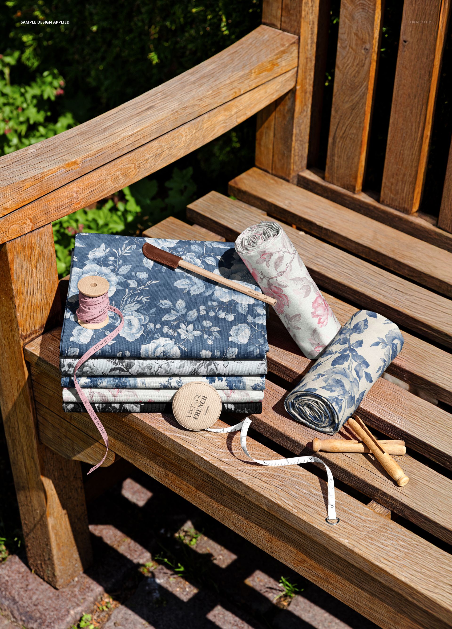 Floral patterned fabric rolls and folded stacks displayed on a rustic wooden bench in a garden setting, PSD mockup.