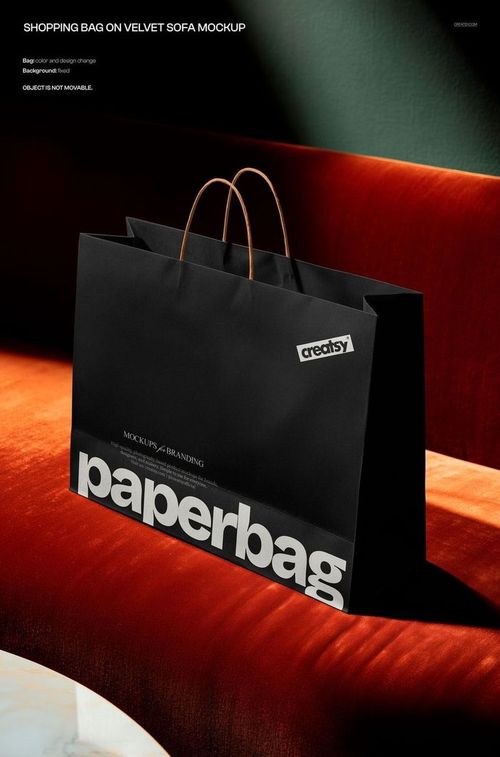 PSD mockup of a black paper shopping bag featuring large white lettering, placed on a deep red velvet sofa.