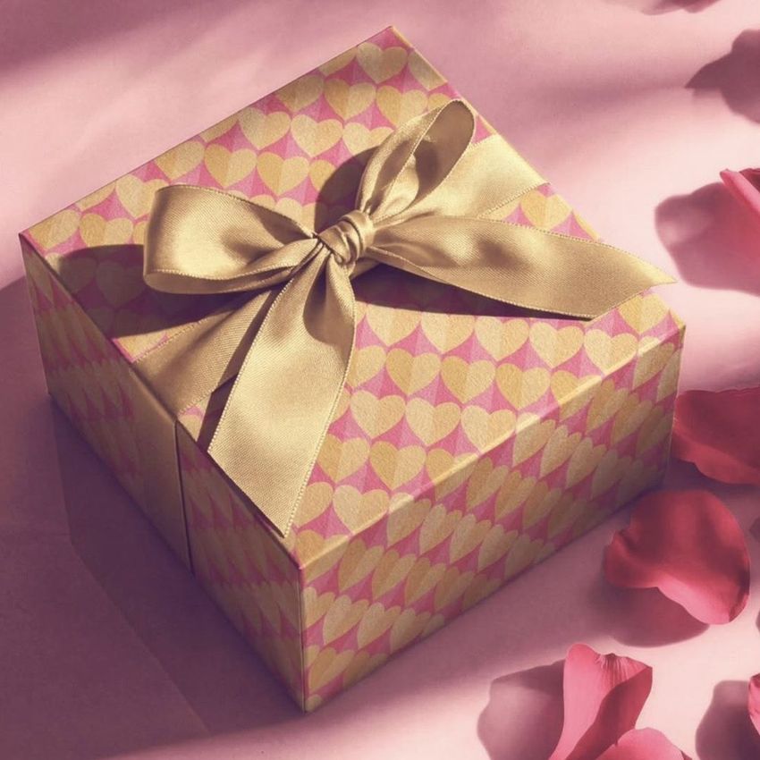 Square gift box mockup with a gold ribbon, covered in a pink and yellow heart pattern, surrounded by scattered rose petals.