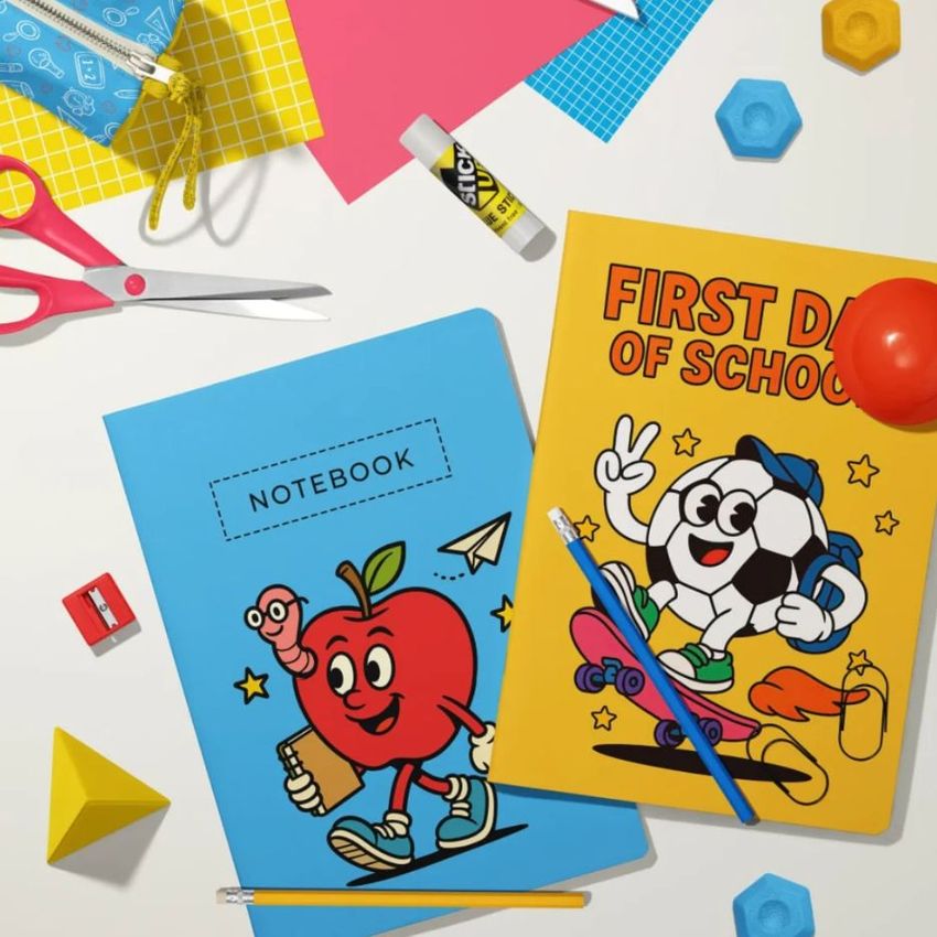 Stationery mockup featuring two classic A4 notebooks on a desk, one blue with an apple illustration and one yellow with a cartoon soccer ball. Various school supplies like scissors, glue stick, colored paper, and pencils are scattered around.