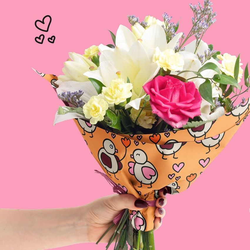 Hand holding a bouquet of mixed flowers wrapped in orange floral packaging with cartoon birds and hearts, against a pink background.