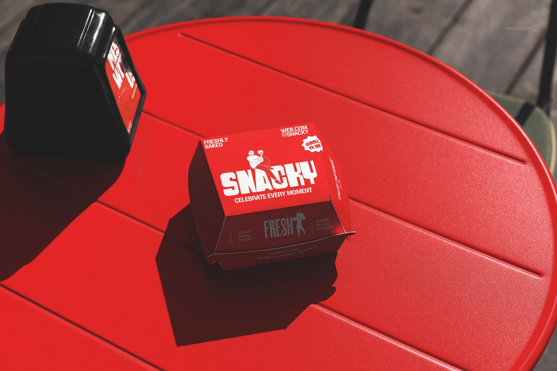 Fast food burger packaging mockup featuring a red box with bold white text on a red table in a sunny outdoor setting.