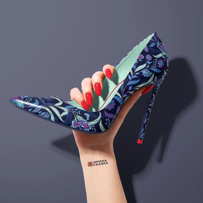 A high heel shoe mockup with a dark blue base and colorful floral pattern, held in a hand with red nails against a gray background.