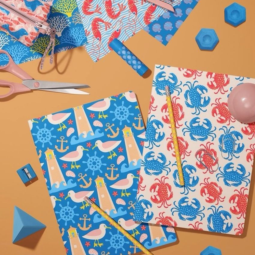 Stationery mockup featuring two classic A4 notebooks with nautical-themed covers, surrounded by pencils, scissors, and geometric props on a tan surface.