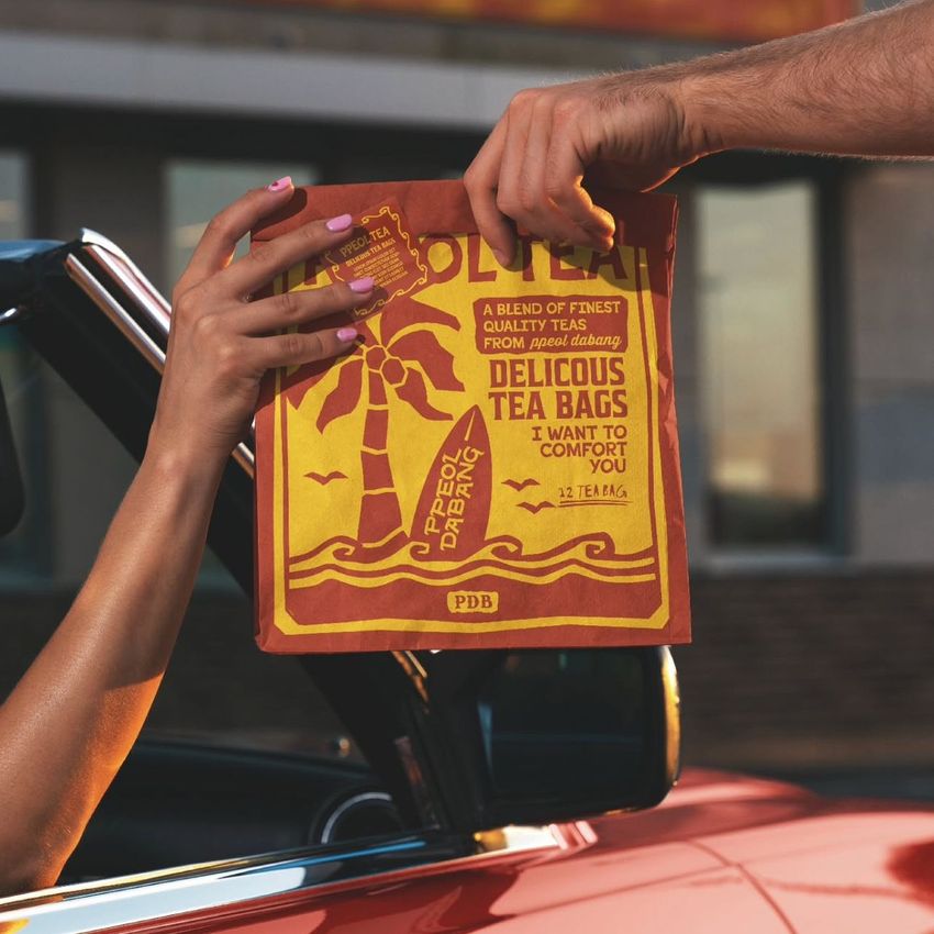 A person hands a printed fast food bag with bold yellow and red graphics through a car window at a drive-thru, with text visible.