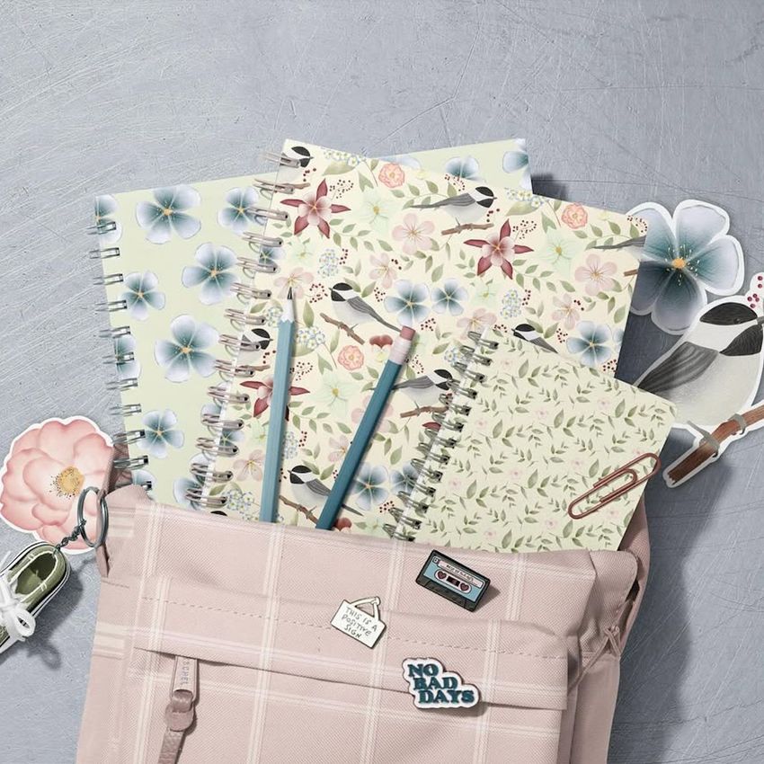 School backpack mockup with spiral notebooks featuring floral and bird patterns, pastel pencils, and decorative pins on a gray surface.