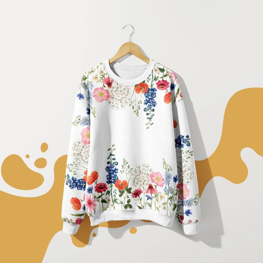 Sweatshirt mockup featuring a white adult pullover with a colorful floral pattern, hanging on a wooden hanger against a white wall with mustard yellow abstract shapes.