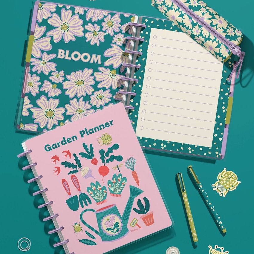 Top view of two spiral planners with floral and gardening designs, surrounded by pens, a pencil case, and stickers on a teal background.