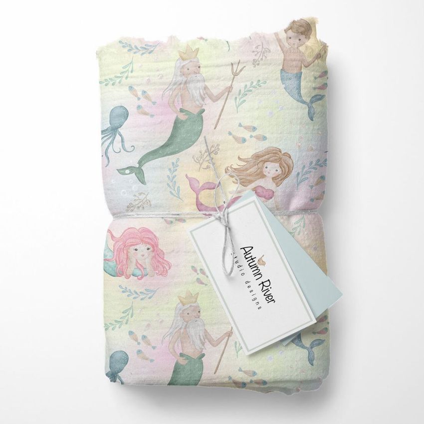 Muslin swaddle blanket mockup featuring a pastel mermaid and sea creature pattern, folded with a branded tag attached.