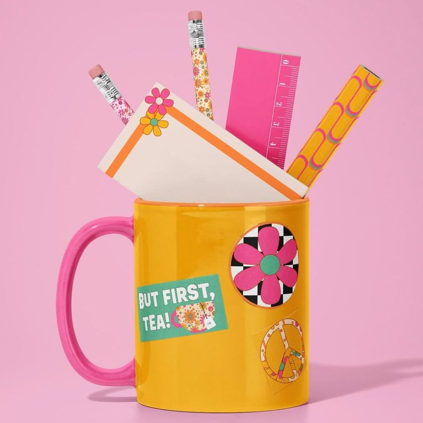 Mug mockup featuring a yellow mug with pink handle, decorated with colorful stickers and filled with stationery items on a pink background.