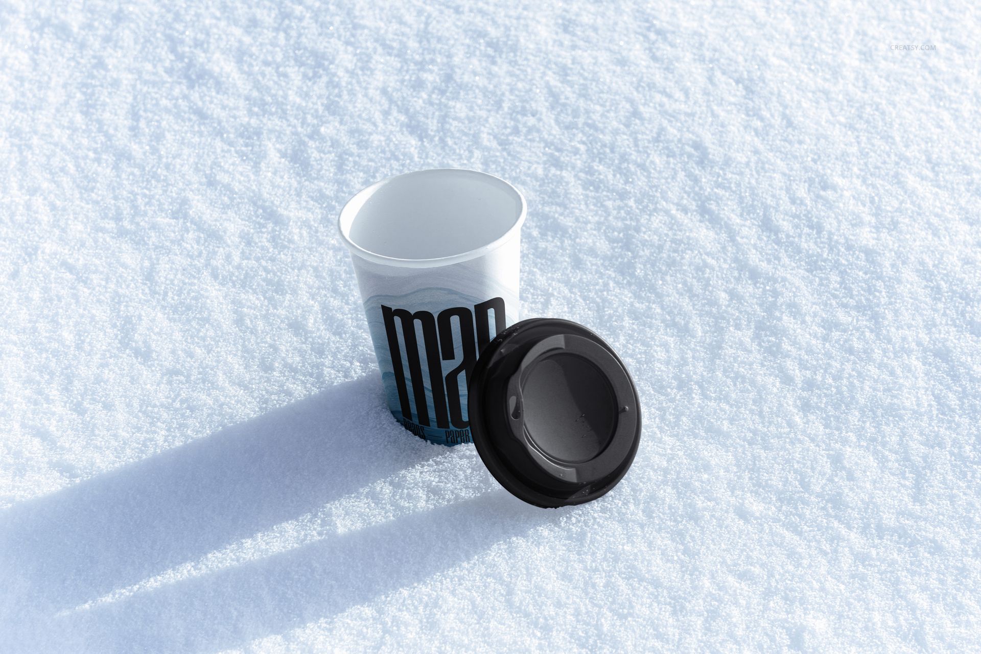 PSD mockup of a white paper cup with black lid on textured snow, viewed from above with sunlight casting shadows.