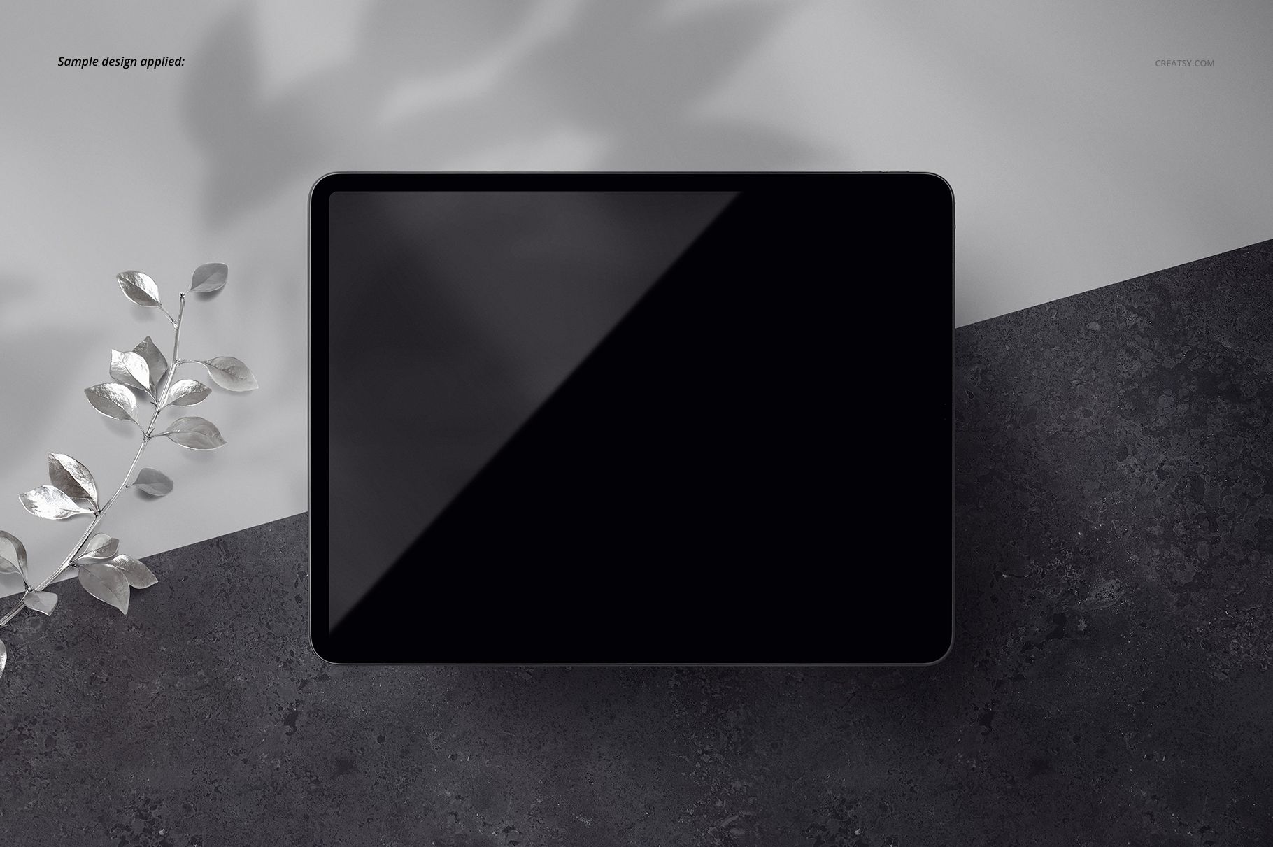 Top view of a black iPad Pro tablet mockup on a dark textured surface with a branch of silver leaves nearby.