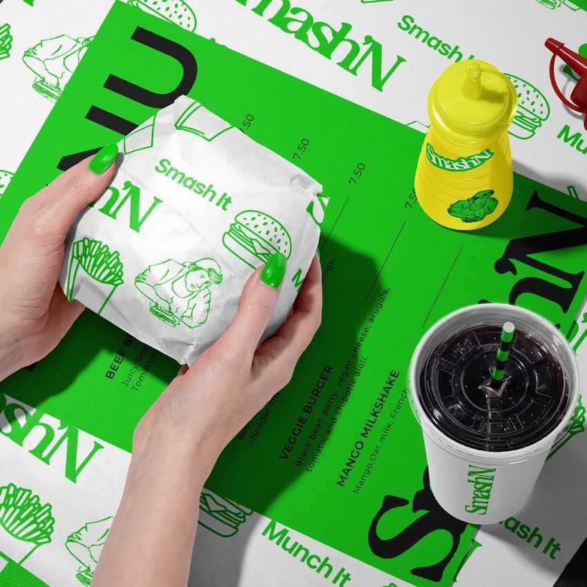 Burger packaging mockup with green and white wrappers, a printed menu, a drink cup, and a yellow sauce bottle on a patterned surface.