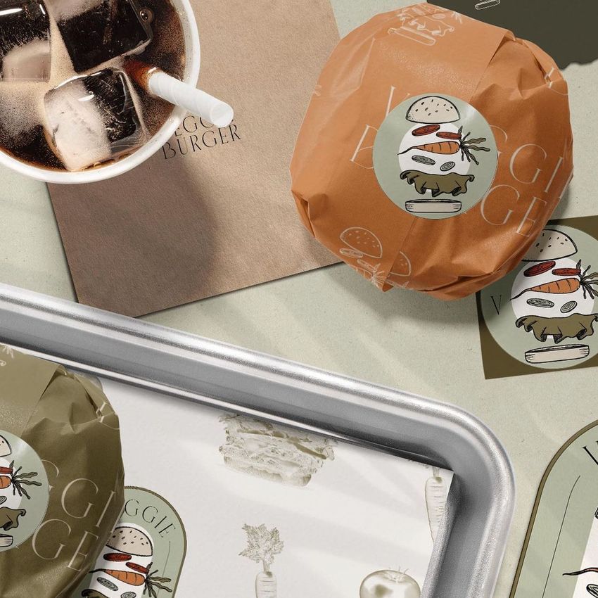 Burger packaging mockup featuring a wrapped veggie burger, a drink with ice, and a napkin with text on a light surface.